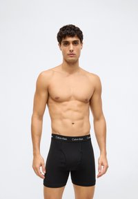 Muž stojící čelem vpřed v černých boxerkách Calvin Klein na světlém jednobarevném pozadí.