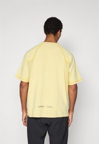 Light yellow athletic t-shirt with short sleeves, contrasting yellow stitching, and a logo on the lower back. Features a round neckline and vented design.