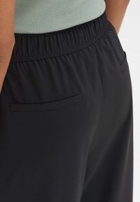 Schwarze, dehnbare Hose mit einem gerafften Bund und dezentem Taschendetail auf der Rückseite. Der Stoff wirkt glatt und hat eine leichte Textur.