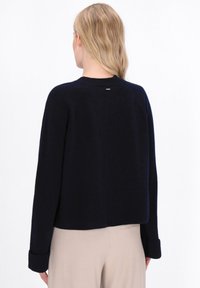 Marineblå cropped sweater med lange ærmer, ribbede manchetter og logo accent på ryggen. Blød tekstur og afslappet pasform, parret med lyse beige bukser.