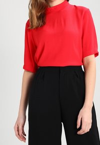 Blouse rouge à manches courtes avec un col haut, en tissu léger, associée à un pantalon noir taille haute avec des plis.