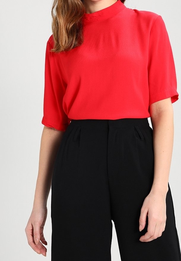 Blouse rouge à manches courtes avec un col haut, en tissu léger, associée à un pantalon noir taille haute avec des plis.