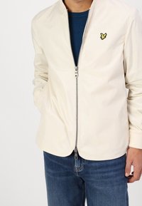 Licht beige jack met een ritssluiting aan de voorkant, voorzien van een gele geborduurde logo. Gestyled met een ronde halslijn en zijzakken. Gecombineerd met blauwe jeans.