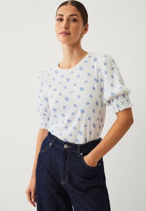 Kvinde med håret samlet bagud iført hvid bluse med blå blomsterprint og puffede ærmer, stoppet ned i mørkeblå højttaljede jeans.