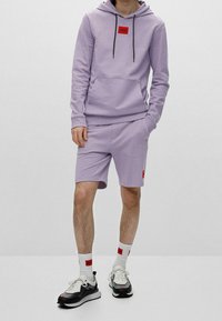 Felpa viola chiaro con lacci neri e logo rosso, abbinata a pantaloni corti coordinati con tasche laterali e un toppe rossa. Calzini bianchi e sneakers.