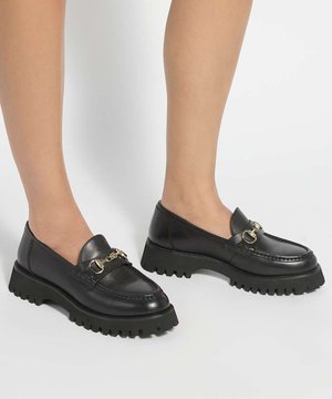 Jambes portant des mocassins en cuir noir à semelle épaisse avec un détail chaîne dorée sur fond blanc.