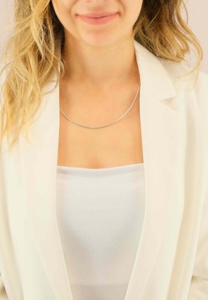 Collana d'argento con un design elegante e curvo, indossata sopra una camicia bianca, completata da un blazer di colore chiaro. Texture liscia, accenti minimalisti.