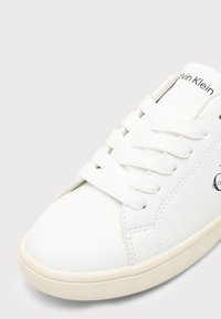 Бял маратонка Calvin Klein с бели дантели и бежова подметка, показваща пришити детайли и частично лого върху езица и страната.