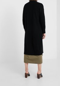 Svart stickad lång cardigan med rak skärning, ribbade manschetter och hög slits bak, ihop med en ljusgrön kjol och svarta mule-skor.