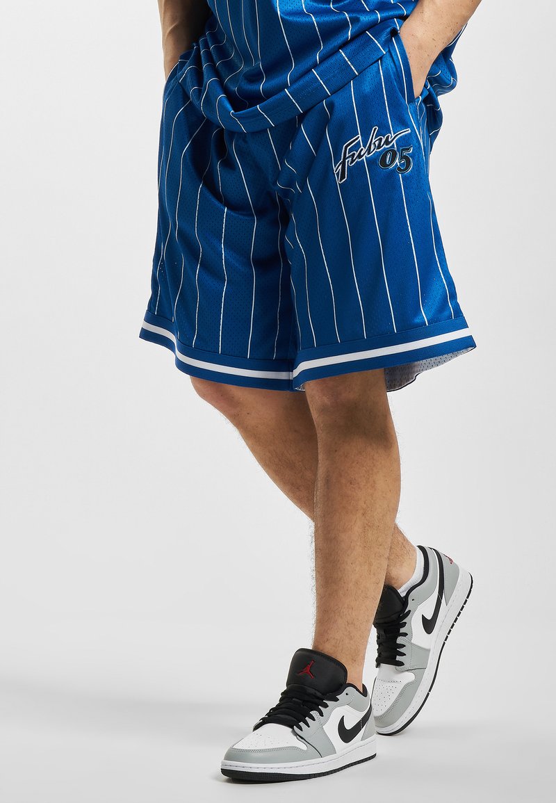 Zilā basketbola šortos ar baltām svītrām, kuru malās ir sānu kabatas un izšūta logo. Kombinēti ar pelēkiem un melniem apaviem.