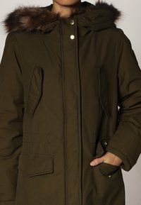 Parka vert olive avec capuche doublée de fausse fourrure, deux poches avant, boutons-pression et fermeture éclair. Présente une texture matelassée.