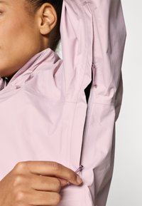 Ανοιχτό ροζ μπουφάν με ψηλό γιακά, φερμουάρ στα πλαϊνά ανοίγματα και διακριτική λάμψη. Διαθέτει θηλιά για κρέμασμα και ρυθμιζόμενο κούμπωμα.