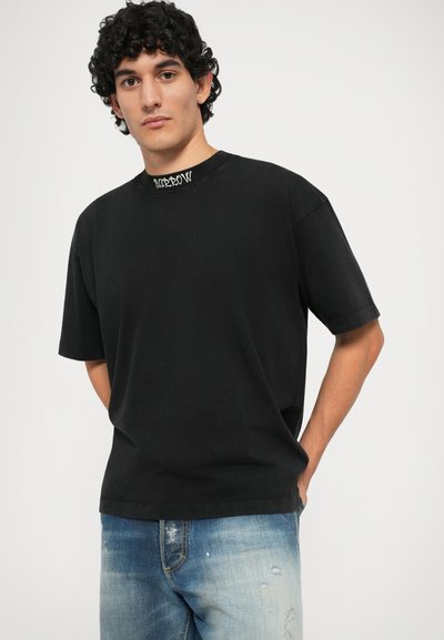 Svart bomull t-skjorte med korte ermer og rund hals. Har en hvit grafisk detalj ved kanten. Båret sammen med lyseblå slitne jeans.