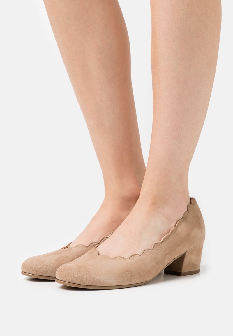 Femme portant des chaussures à talon bas en daim beige avec des bords en forme de vagues, se tenant devant un fond blanc uni.