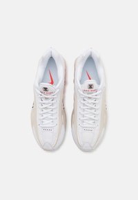 Paire de baskets blanches Nike Shox avec des détails latéraux beiges et des logos Swoosh rouges, vues de dessus sur un fond blanc uni.