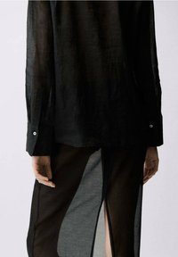 Schwarze, durchsichtige Bluse mit langen Ärmeln und geknöpften Manschetten, in Schichten über einem schwarzen Kleidungsstück mit Seitenschlitzen getragen, das eine glatte Textur aufweist.