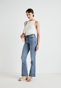 Witte mouwloze top, high-waisted lichtblauwe denim flare jeans en lichtbruine sandalen. Model houdt een kleine crossbody tas met een strap vast.