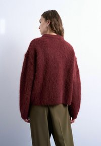 Femme aux cheveux ondulés portant un pull texturé bordeaux et un pantalon vert olive, debout contre un fond clair uni, tournée de dos.