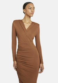 Frau in einem figurbetonten, langärmeligen, rostfarbenen Wickelkleid mit Raffung, die vor einem schlichten weißen Hintergrund steht.