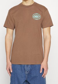 Hnědé bavlněné tričko s krátkým rukávem, se světle modrým logem "OBEY" uvnitř grafiky planety na levé části hrudi.