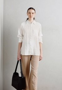 Camicia bianca in lino con bottoni e maniche lunghe, abbinata a pantaloni beige sartoriali. Borsa nera a tracolla. Sfondo grigio chiaro.