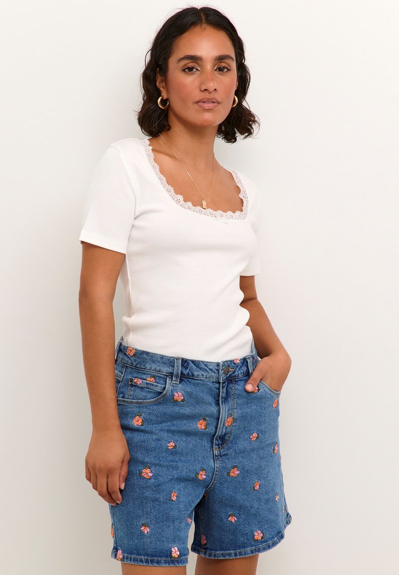 Top blanco de manga corta acanalada con escote cuadrado rematado con encaje, combinado con pantalones cortos de mezclilla azul con bordados florales coloridos.