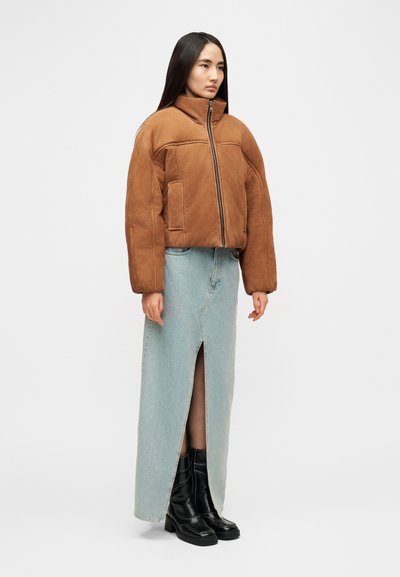 Veste courte marron avec un col montant et une fermeture éclair frontale, associée à une jupe en denim bleu clair avec une fente à l'avant. Bottines noires.