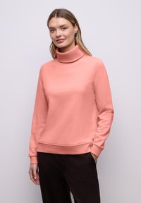 Sweat-shirt couleur pêche avec col montant, manches longues, poignets côtelés et ourlet à bande, porté avec un pantalon foncé. Texture du tissu lisse.