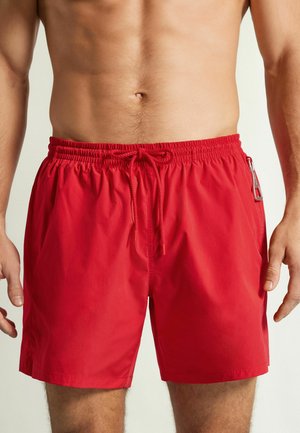 Homme torse nu portant un short de bain rouge à cordon avec une poche latérale zippée, debout devant un fond clair uni.