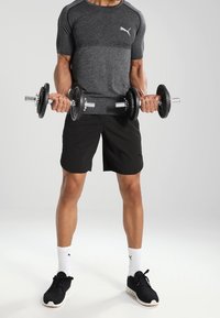 Černé sportovní tričko s šedými detaily, černé kraťasy, držící činky, obuté černé tenisky a bílé ponožky s logem; minimální pozadí.