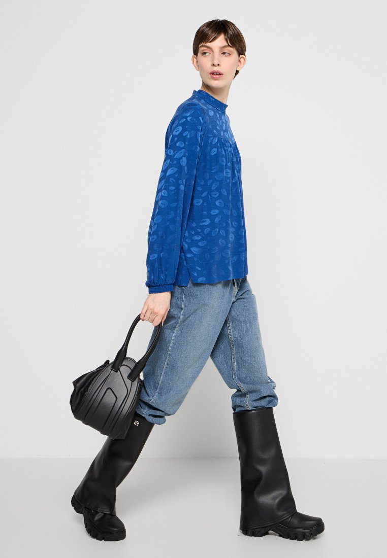 Blaues Langarmblusen mit Blattmuster, kombiniert mit lockeren Denim-Jeans und schwarzen knielangen Gummistiefeln. Schwarze Handtasche mit strukturiertem Design.