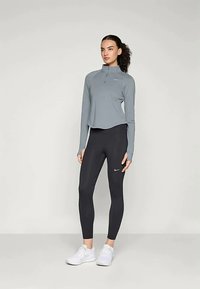 Szürke, hosszú ujjú sportos felső negyed cipzárral és logóval, fekete, testhezálló leggings-szel, amelyen szintén van logó, valamint fehér sportcipővel párosítva.