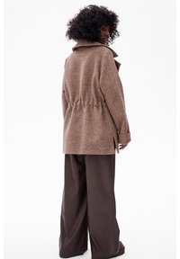 Bruine wolmix jas met een brede kraag, taaige taille en zijsplits; gecombineerd met hooggewaisted donkerbruine neplederen broek.