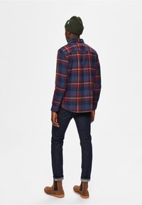 Rutig flanellskjorta i röd, blå och vit toner; har en knapp-ner krage och rak rygg. Bärs med mörka jeans och bruna chelsea-boots.