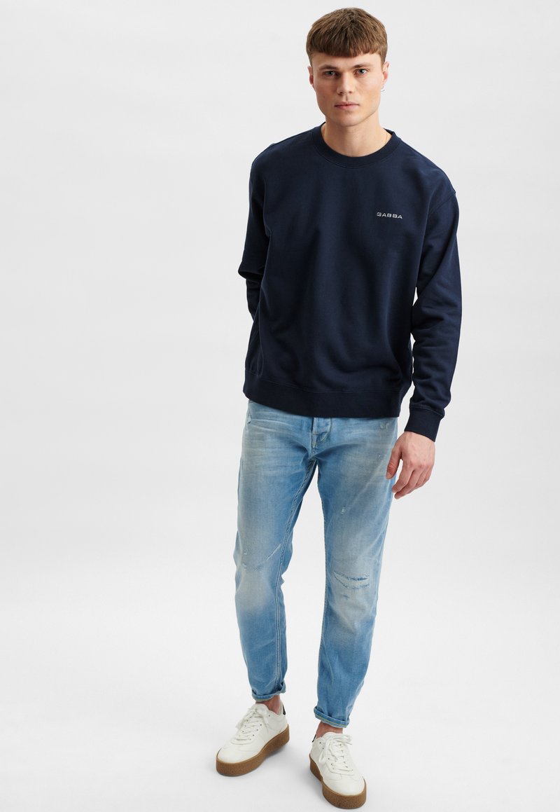 Marineblauwe sweatshirt met ronde hals en geribbelde manchetten, gecombineerd met lichtblauwe versleten jeans en witte sneakers met gestructureerde zolen.