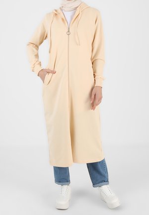 Kvinde iført beige lang hætteklædedragt med lynlås, lyseblå op-rullede jeans, hvide sneakers og beige tørklæde, stående med den ene hånd i lommen.