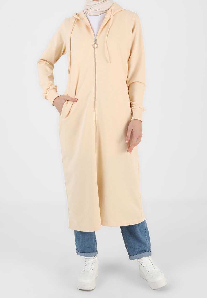 Femme portant une longue robe à capuche beige avec fermeture éclair, un jean bleu clair retroussé, des baskets blanches et un foulard beige, debout avec une main dans la poche.