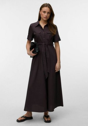 Vrouw draagt een donkerbruine korte mouwen overhemdjurk met een ceintuur in de taille, houdt een zwarte clutch vast en draagt zwarte sandalen.