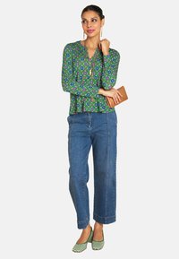 Grün gemusterte Bluse mit langen Ärmeln, kombiniert mit hoch taillierten blauen Jeans. Grün-weiß gestreifte Schuhe, Halten einer sandfarbenen Clutch.