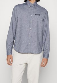 Košile s knoflíky vyrobená z texturované modré tkaniny, s límečkem, dlouhými rukávy a malým vyšitým logem "BOSS" na hrudi.
