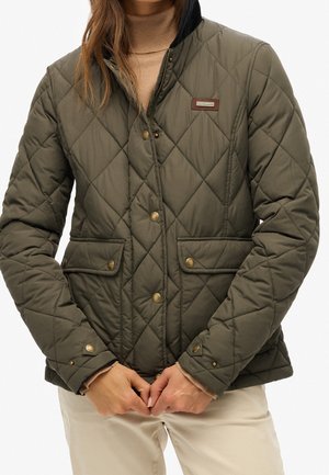 Kobieta w oliwkowo-zielonej pikowanej kurtce z zapięciem na guziki i przednimi kieszeniami z klapkami, nosząca beżowy golf i jasne spodnie.