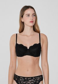 Černá krajková bralette s vlnitými okraji, s podšívkou na podporu, tenkými nastavovacími ramínky a odpovídajícími krajkovými kalhotkami. Hladký povrch, jemný design.