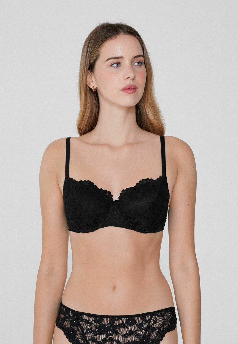 Černá krajková bralette s vlnitými okraji, s podšívkou na podporu, tenkými nastavovacími ramínky a odpovídajícími krajkovými kalhotkami. Hladký povrch, jemný design.