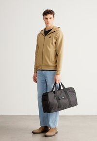 Čierna taška typu duffel s dvoma rukoväťami, hladkou textúrou a jemným logom, doplnená béžovou mikinou na zapínanie a svetlomodrými džínsami.