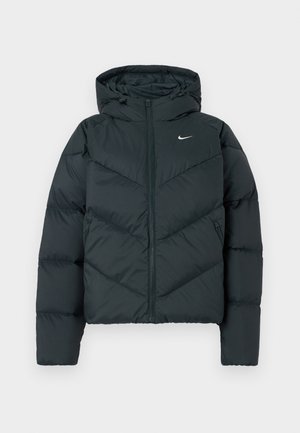 Dunkelgrüne Pufferjacke mit Kapuze, mit einem Reißverschluss, Chevron-Steppnähten und einem kleinen weißen Logo auf der linken Brust.