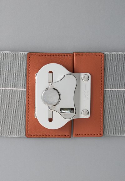 Gros plan d’une boucle de sécurité métallique argentée attachée à une sangle grise avec des patchs en cuir marron sur un fond en tissu gris.