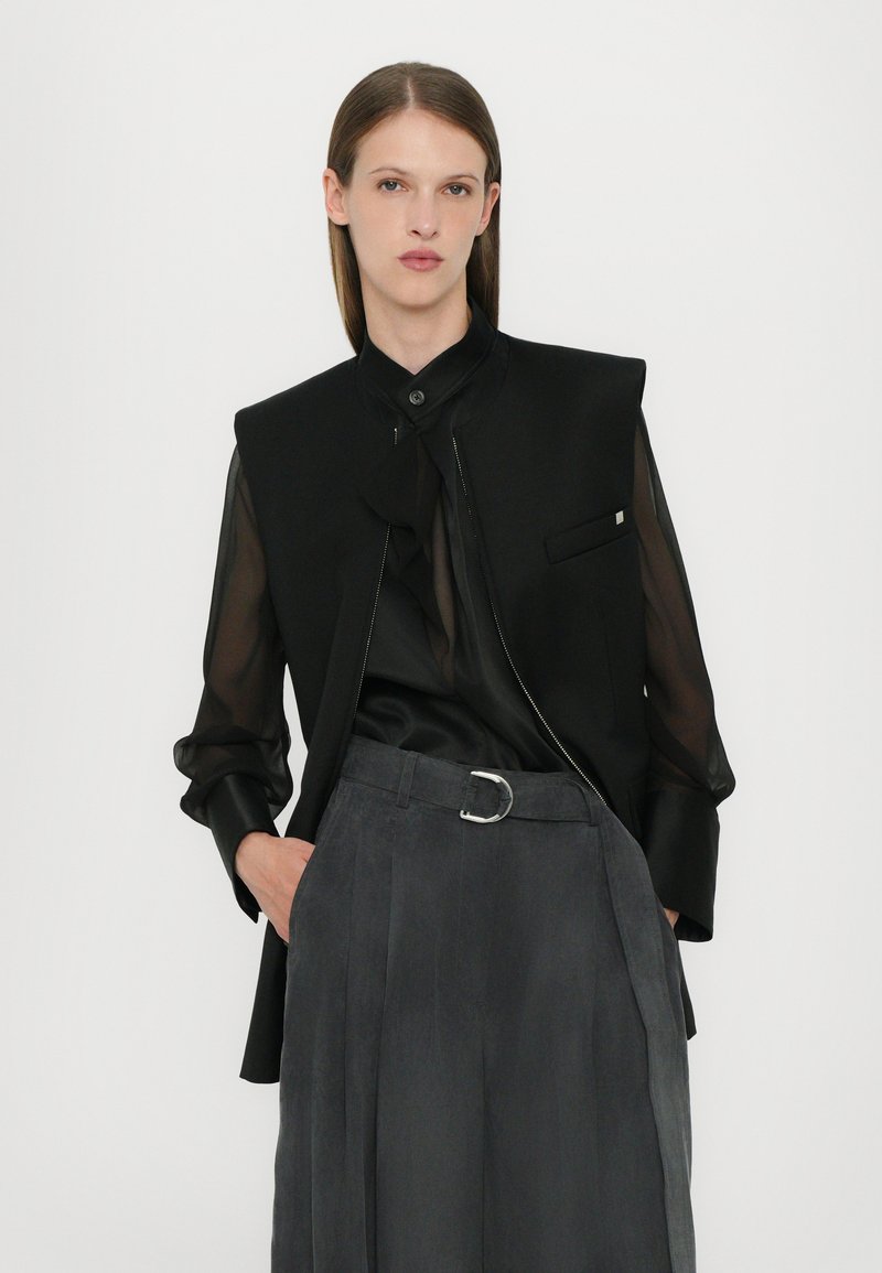 Black sleeveless blazer with a high neckline and zip front, paired with a black semi-sheer shirt and gray belted trousers.