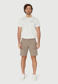 Pantalones cortos de carga beige cortos con dos bolsillos laterales, combinados con una camiseta blanca de cuello redondo que presenta detalles del logo en negro. Llevado con zapatillas blancas.