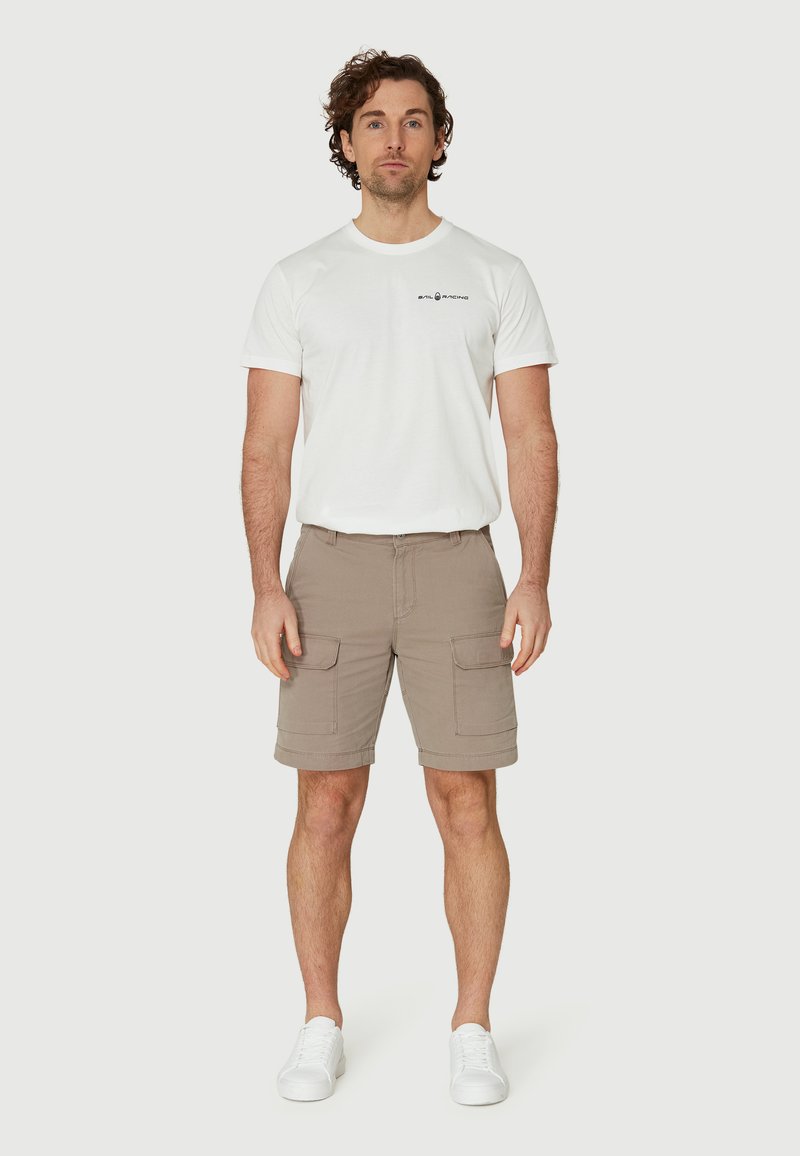 Pantalones cortos de carga beige cortos con dos bolsillos laterales, combinados con una camiseta blanca de cuello redondo que presenta detalles del logo en negro. Llevado con zapatillas blancas.