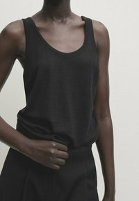 Femme portant un débardeur noir sans manches et un pantalon noir, avec une main serrant le haut près de la taille, sur un fond uni.